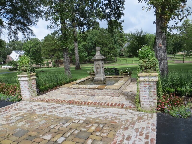 Hooglands Water Features Fountains Waterfalls Ponds – 01 Elegant stone fountain surrounded by lush greenery and brick pathway, enhancing outdoor landscape aesthetics for tranquil retreats.