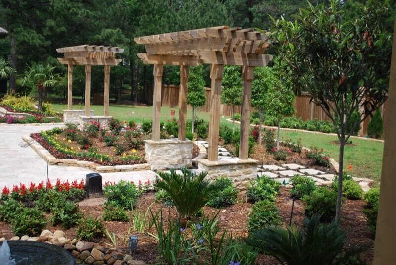 hooglands Pergolas Gazebos Arbors - 01 Custom wooden pergolas surrounded by vibrant flower beds and lush greenery in a landscaped yard, enhancing outdoor elegance and providing shaded areas.