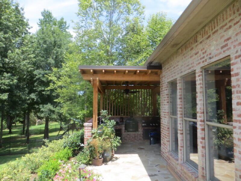 hooglands Pergolas Gazebos Arbors - 03 Custom wooden pergola attached to a brick home, surrounded by lush greenery and flowering plants, enhancing outdoor living space.